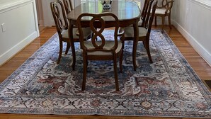 A dining room with a round table, six chairs, a chandelier, and a large patterned rug.