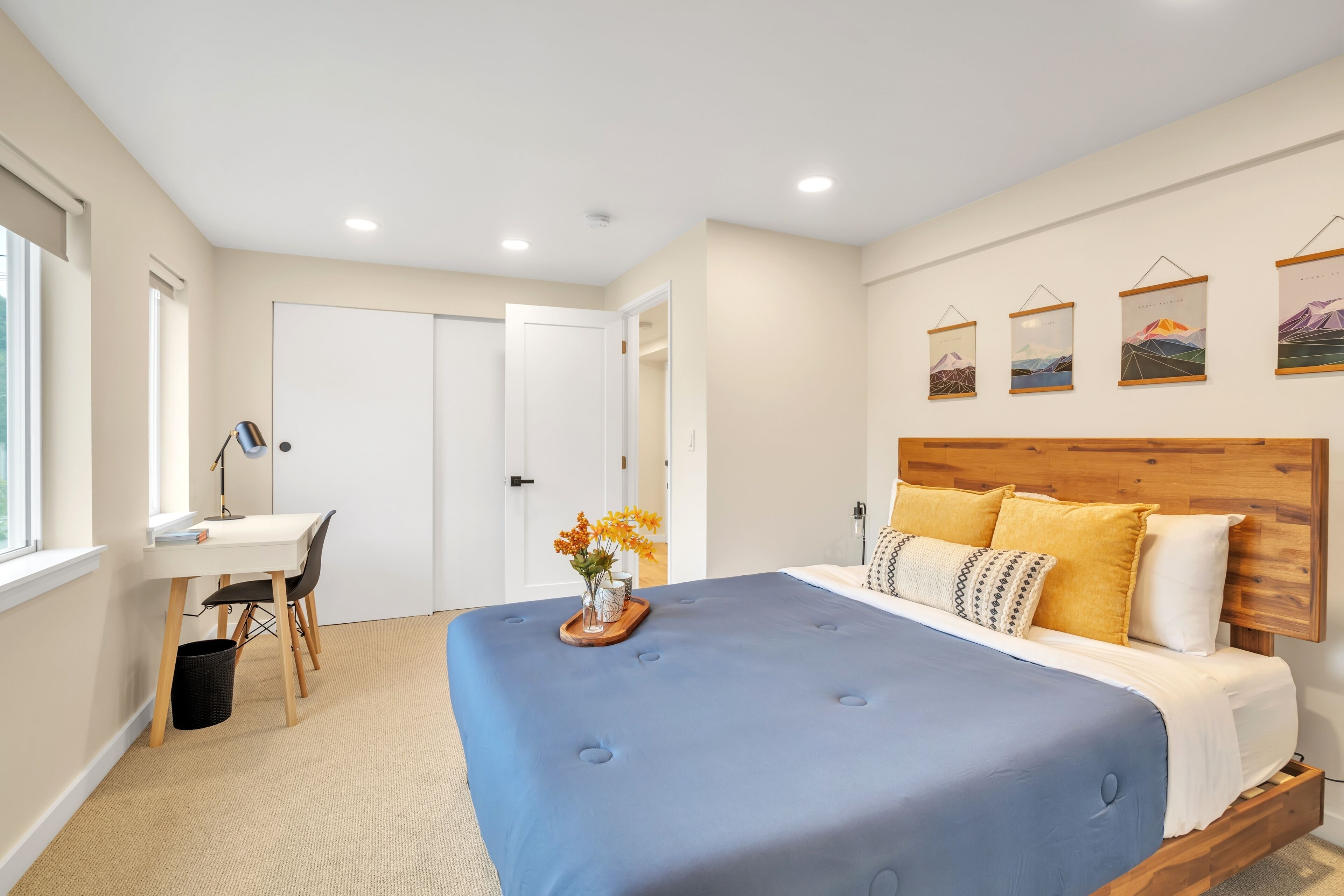 A bedroom with a large bed, a desk, and a chair. There are wall art pieces above the bed and a door leading to another room.