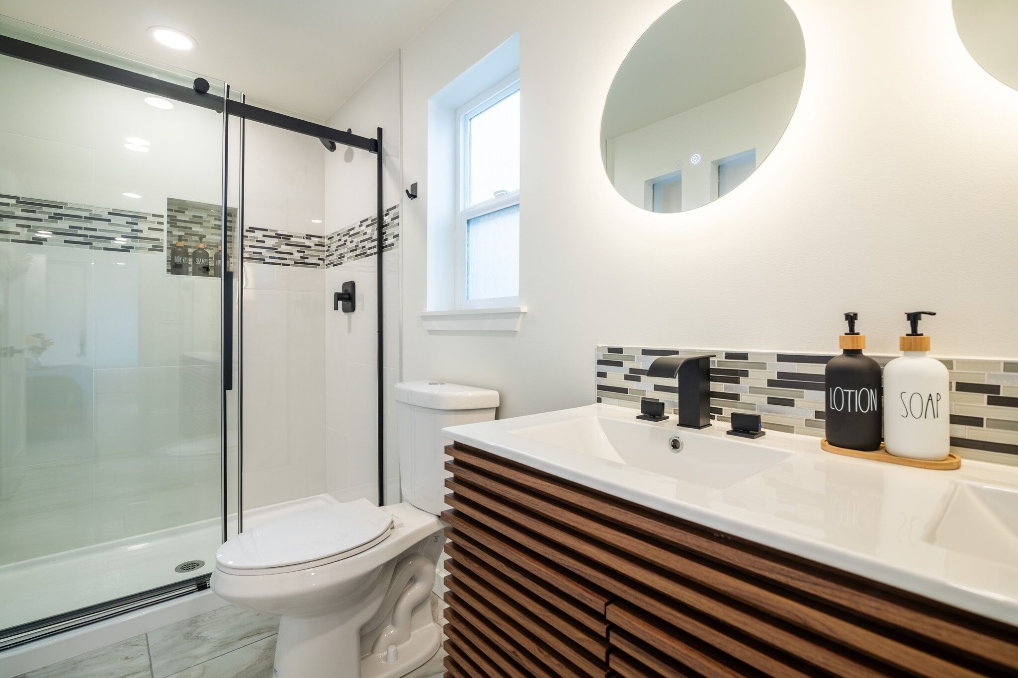 Une salle de bain moderne dotée d’une douche à parois de verre, de toilettes, d’un meuble-lavabo avec un motif en panneaux de bois et d’un miroir rond.