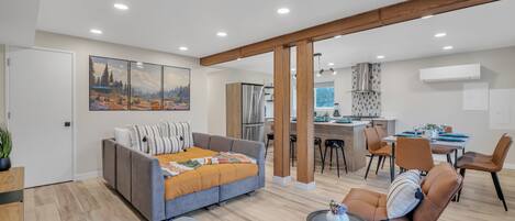 A modern living room with a grey sofa, a blue ottoman, and a dining area with brown chairs and a table.