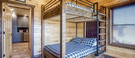 A bunk bed room with a kitchen area, wooden walls, and a window with blinds.