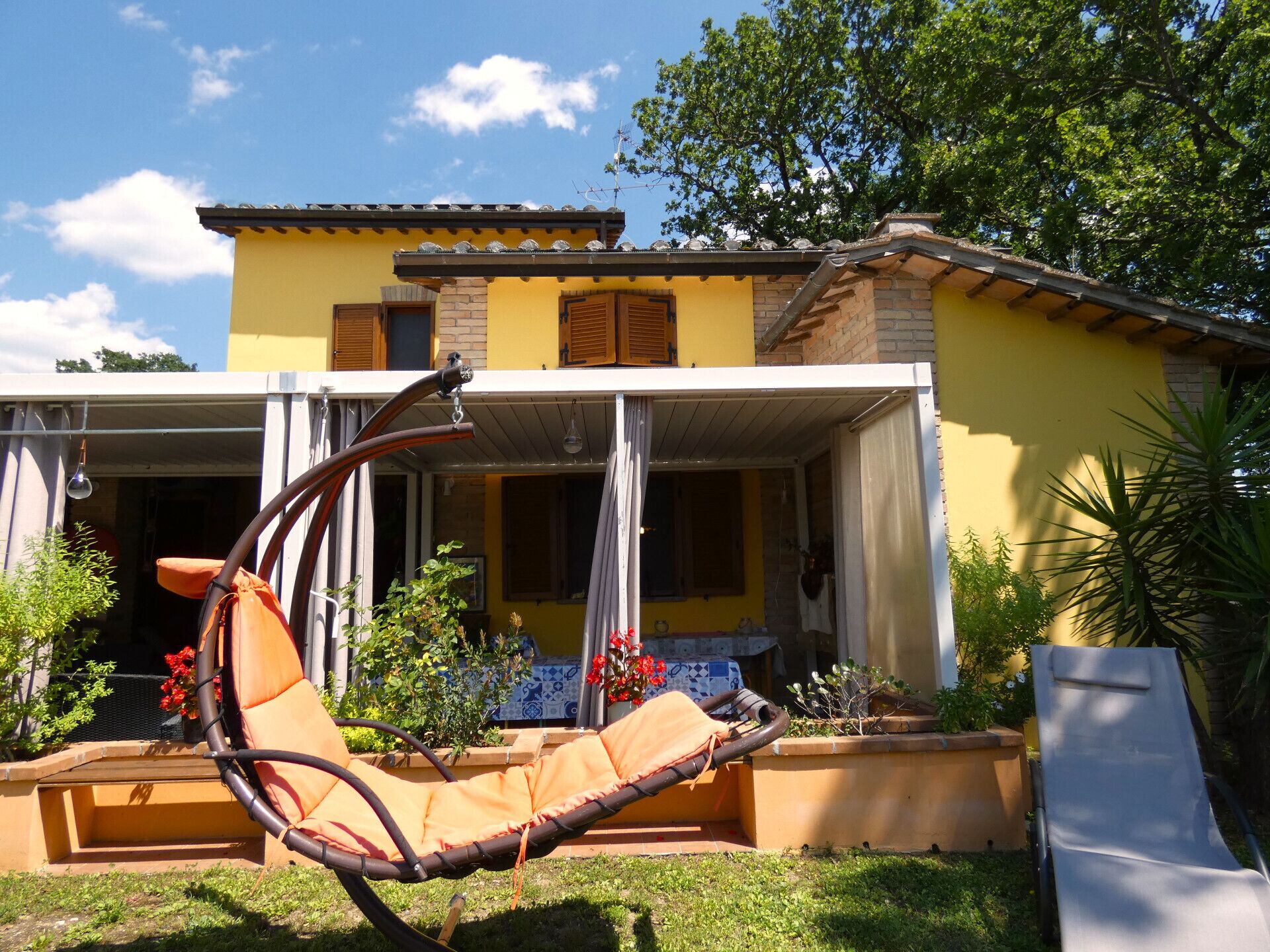 A yellow house with a covered porch, a hanging chair, and a garden with potted plants and flowers.