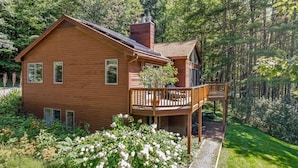 A two-story wooden house with a balcony, surrounded by lush greenery and trees.