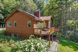 A two-story wooden house with a balcony, surrounded by lush greenery and trees. - Highland Ridge House with Game Room & Large Deck (Quechee)