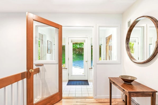 A hallway with a wooden door leading to a room, a round mirror on the wall, and a wooden table with a bowl.