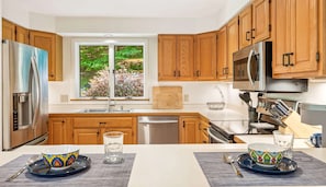 A modern kitchen with wooden cabinets, stainless steel appliances, and a window with a view of greenery.