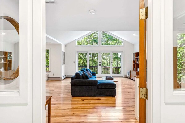A spacious living room with a large window, a dark sofa, and a wooden floor.