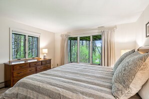 A bedroom with a large bed, a wooden dresser, and a window with a forest view.