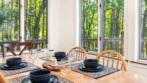 A dining room with a table set for four, large windows with a forest view, and a wooden chair.