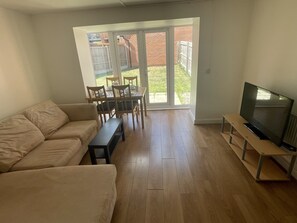 Ein Wohnzimmer mit einer Couch, einem Fernseher auf einem Schrank, einem Esstisch mit Stühlen und einer Glasschiebetür, die zu einem Garten führt.