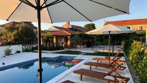 Un espace piscine doté de chaises longues et de parasols, entouré d’un jardin et de bâtiments.