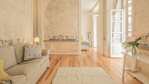 Een kamer met een houten vloer, een beige bank, een witte vloerkleden en een bed met een blauw hoofdeinde.