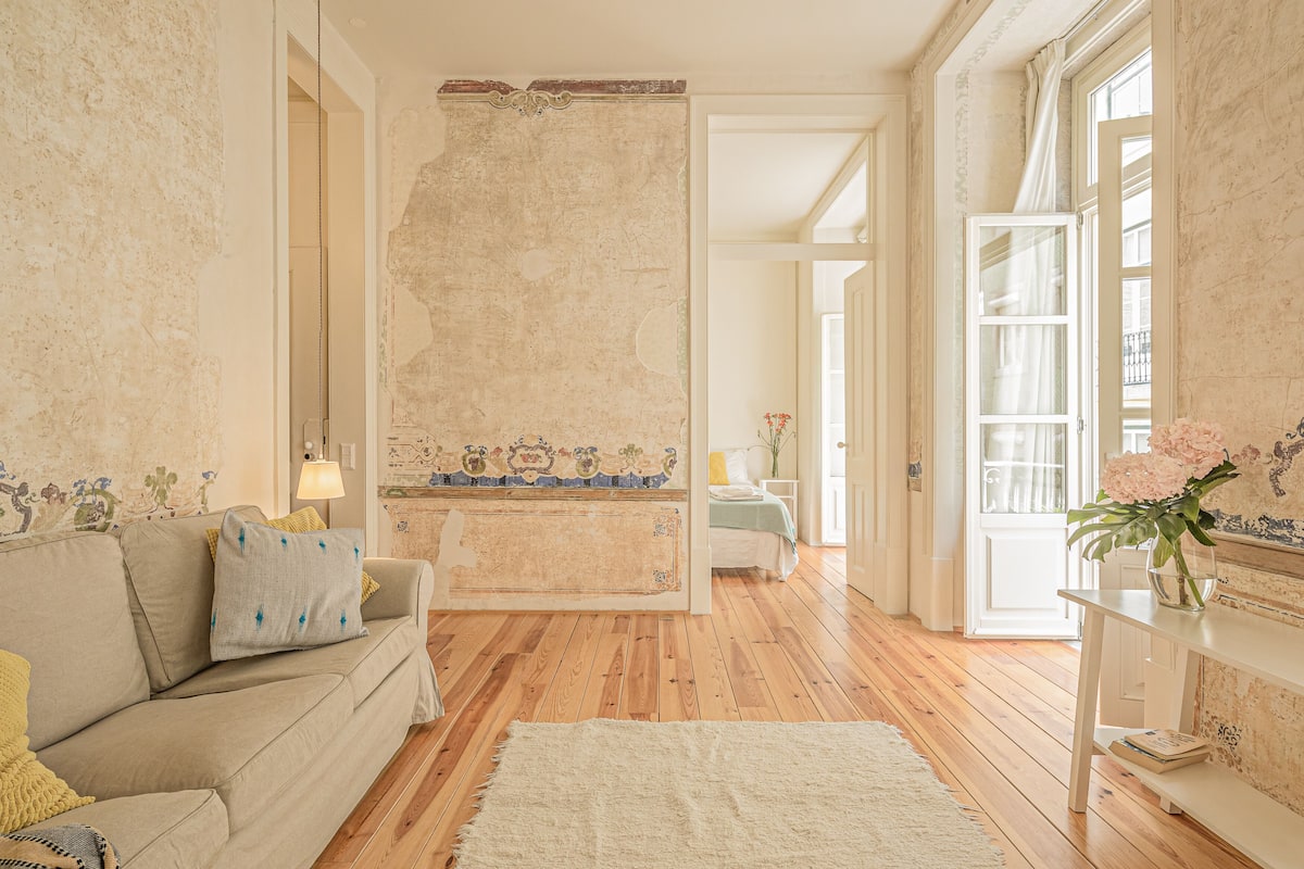Een kamer met een houten vloer, een beige bank, een witte vloerkleden en een bed met een blauw hoofdeinde.