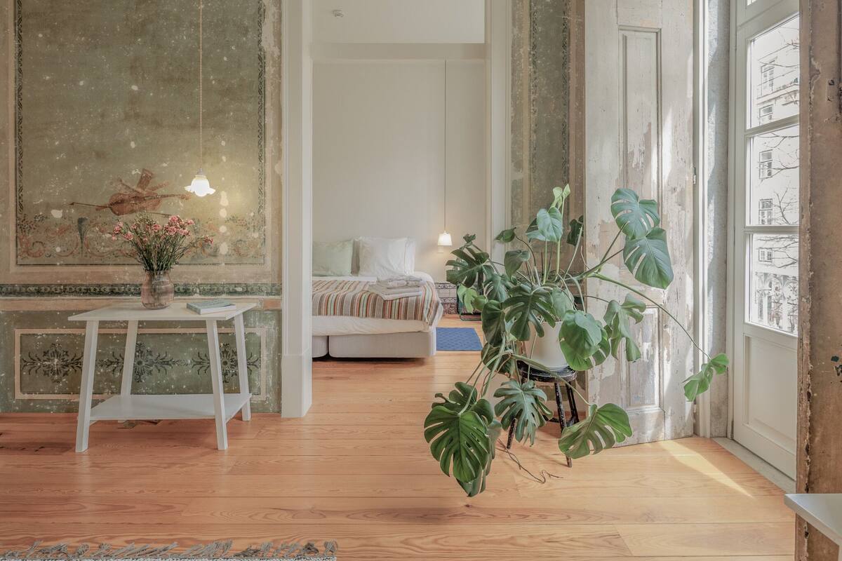 Een kamer met een houten vloer, een bed met gestreepte beddengoed, een tafel met een vaas bloemen en een grote plant.