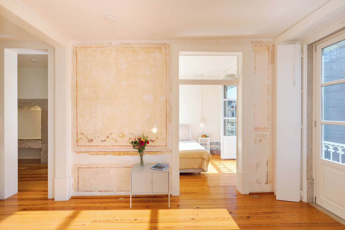 Een kamer met houten vloer, een bed, een nachtkastje met een bloevas en een groot raam.