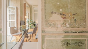 Een eethoek met een glazen tafel en stoelen, een uitzicht op de stad door grote ramen, en een gedeeltelijk gerestaureerde muur met een gitaar en bloemendecoratie.