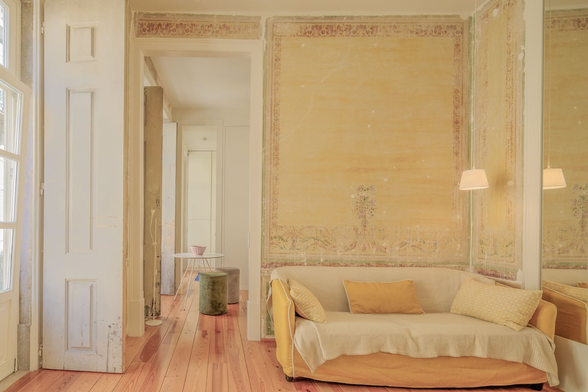 Een kamer met een houten vloer, een beige bank met kussens, een klein rond tafeltje en een spiegel met een lamp.