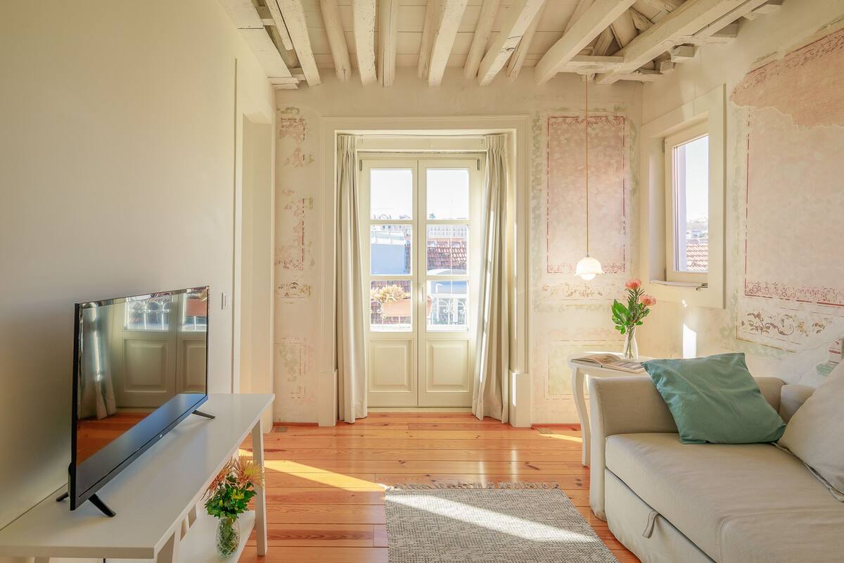 Een woonkamer met een televisie op een witte kast, een bank met een groen kussen, een kleine tafel met een lamp en een deur die naar buiten leidt.