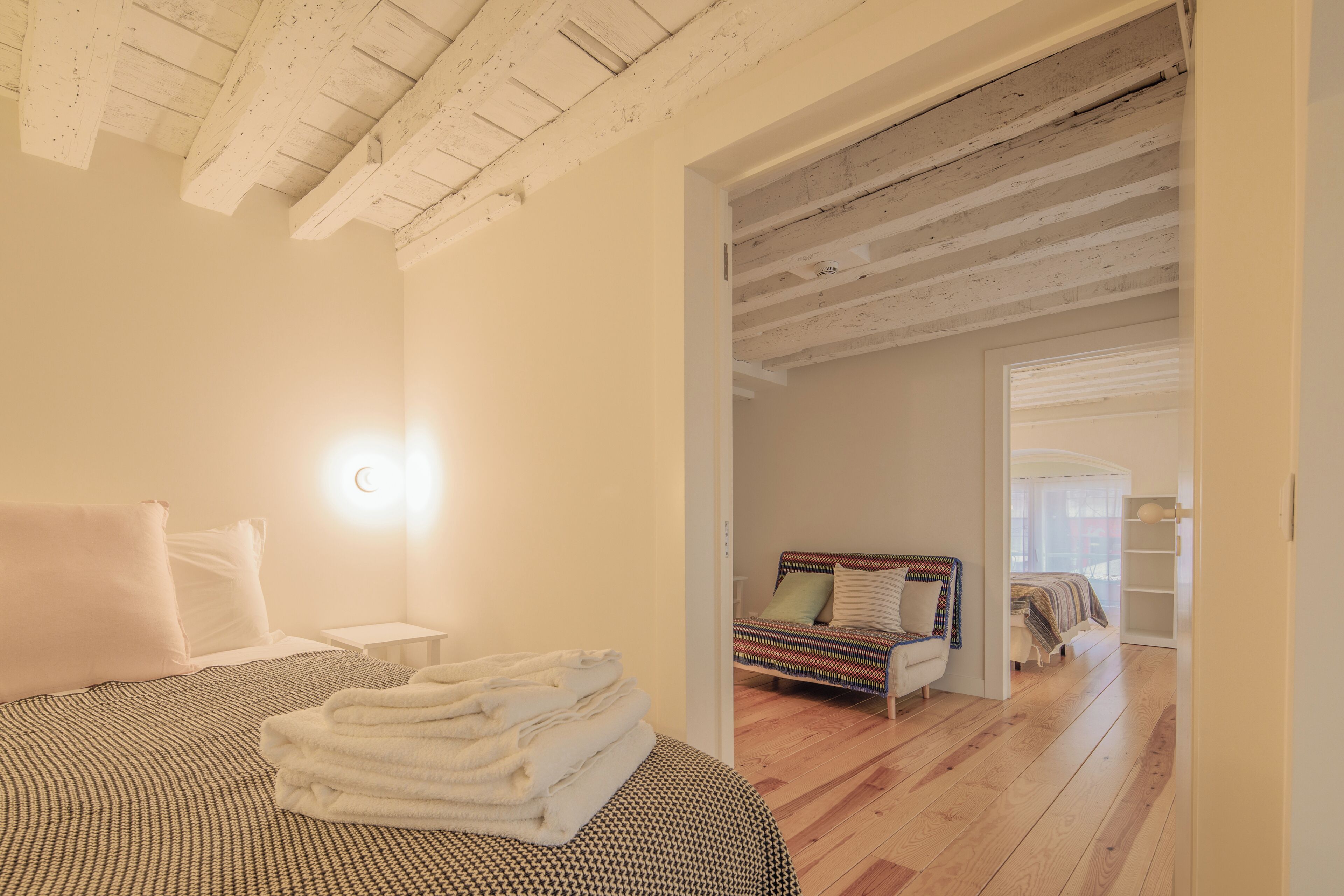 A bedroom with a bed, a bedside table, a lamp, and a view into another room.