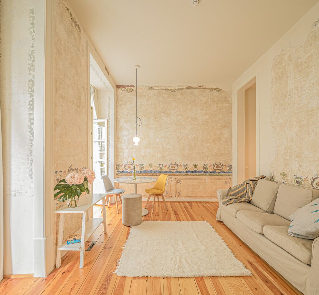 Sala de estar con piso de madera, un sofá color beige, alfombra blanca, una pequeña mesa redonda con sillas y un jarrón con flores.