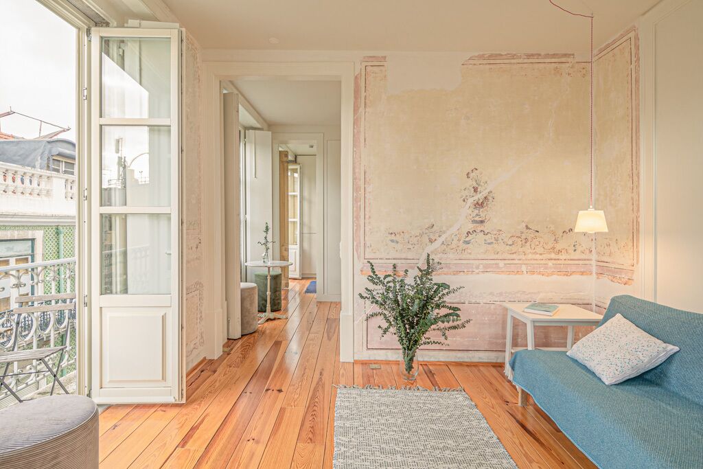Una habitación con piso de madera, un sofá azul, una mesita con una lámpara, una planta y vistas a través de una puerta abierta.