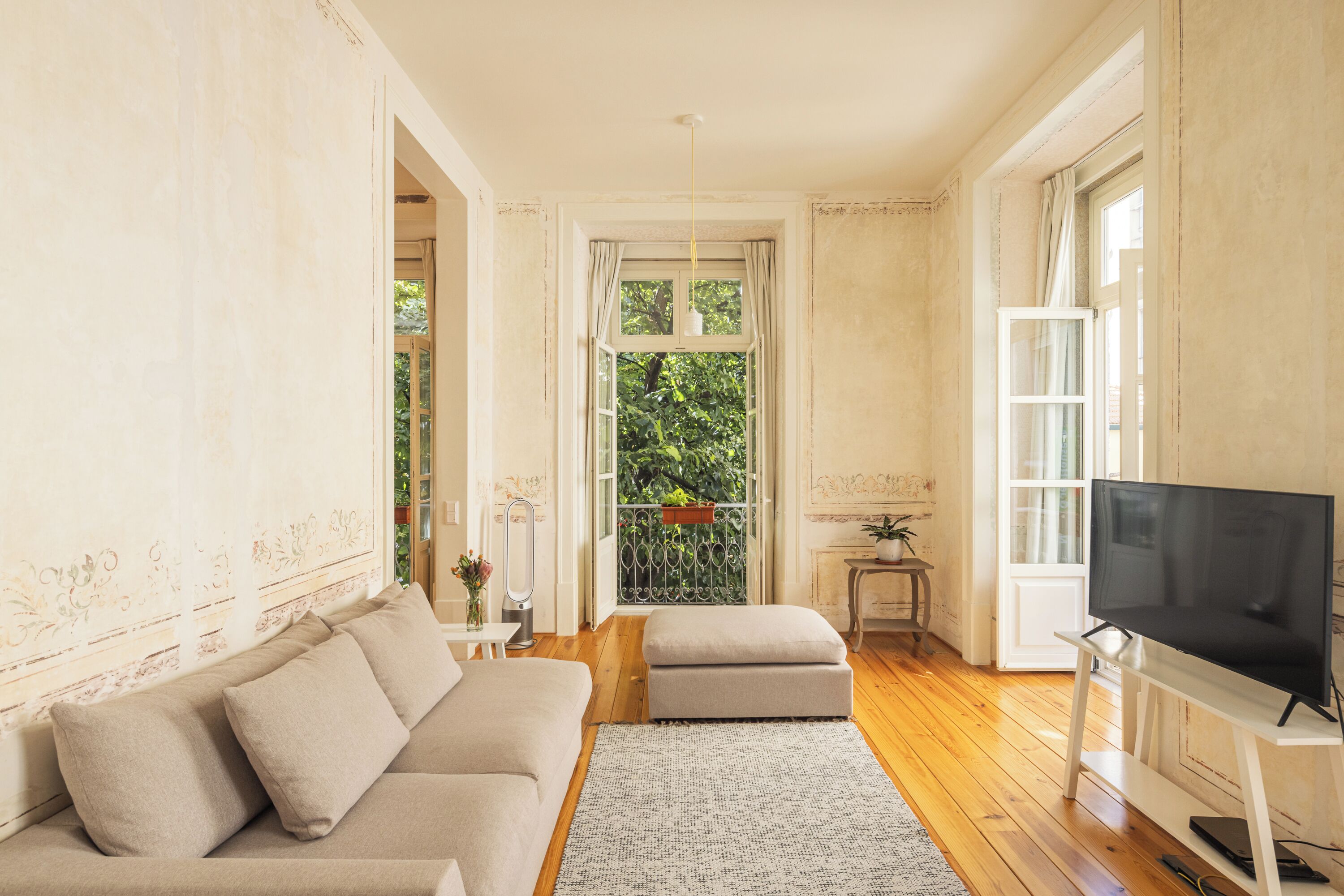 A living room with a sofa, ottoman, TV, and a view of greenery outside.