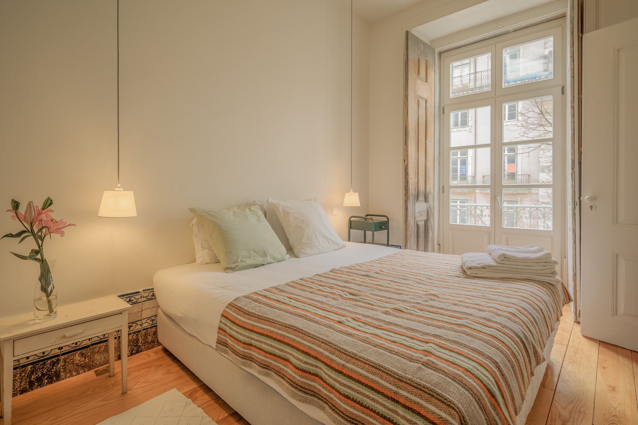 Un dormitorio con cama, lámparas de noche, mesita de noche, un jarrón con flores y ventana con vista a edificios.
