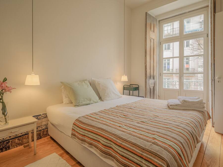 Un dormitorio con cama, lámparas de noche, mesita de noche, un jarrón con flores y ventana con vista a edificios.