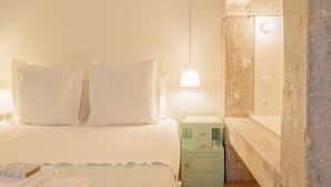 Een slaapkamer met een bed, kussens, een nachtkastje, een lamp en een stenen muur.