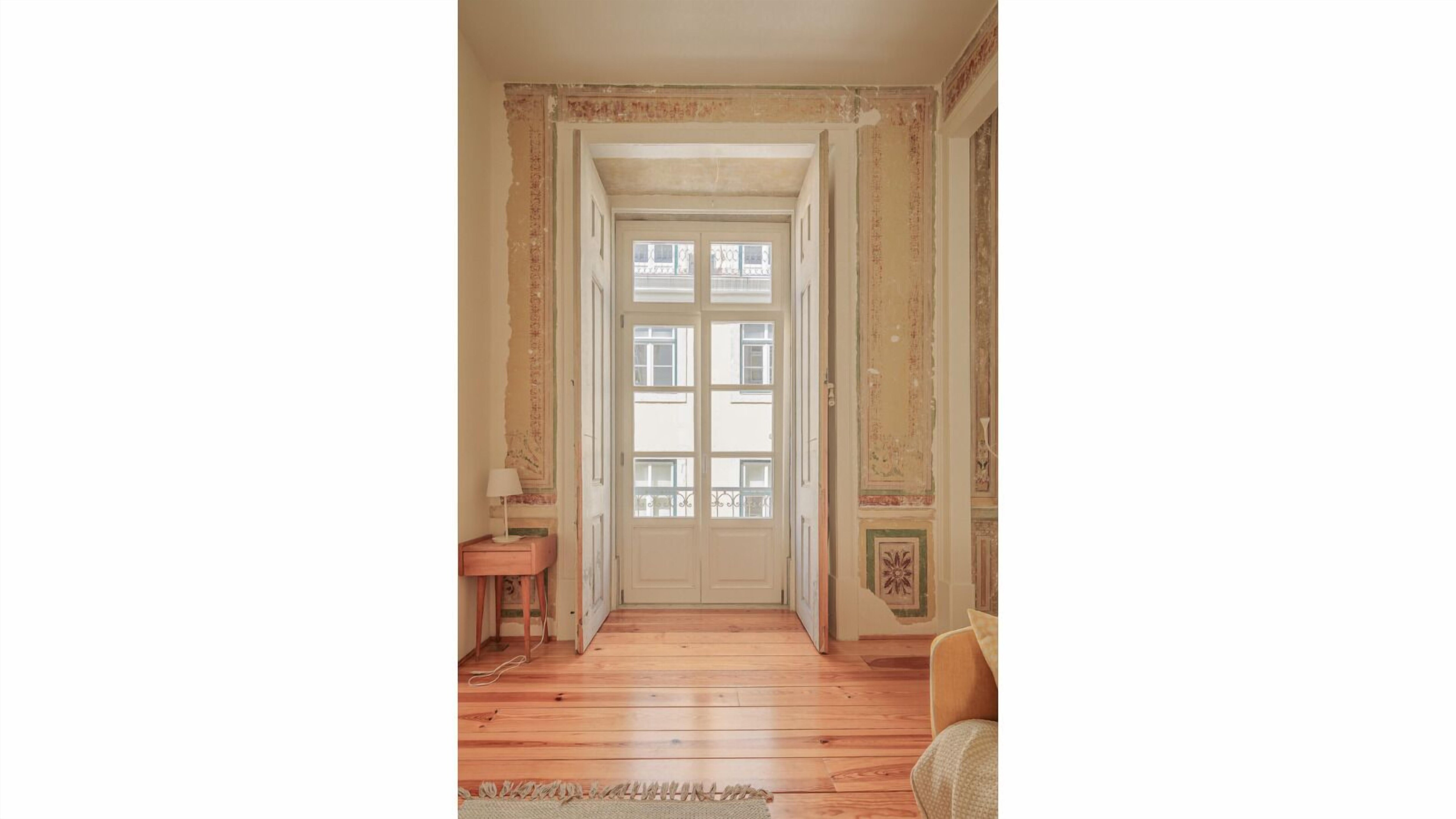 Um corredor com piso de madeira, uma porta com painéis de vidro e uma pequena mesa com abajur.