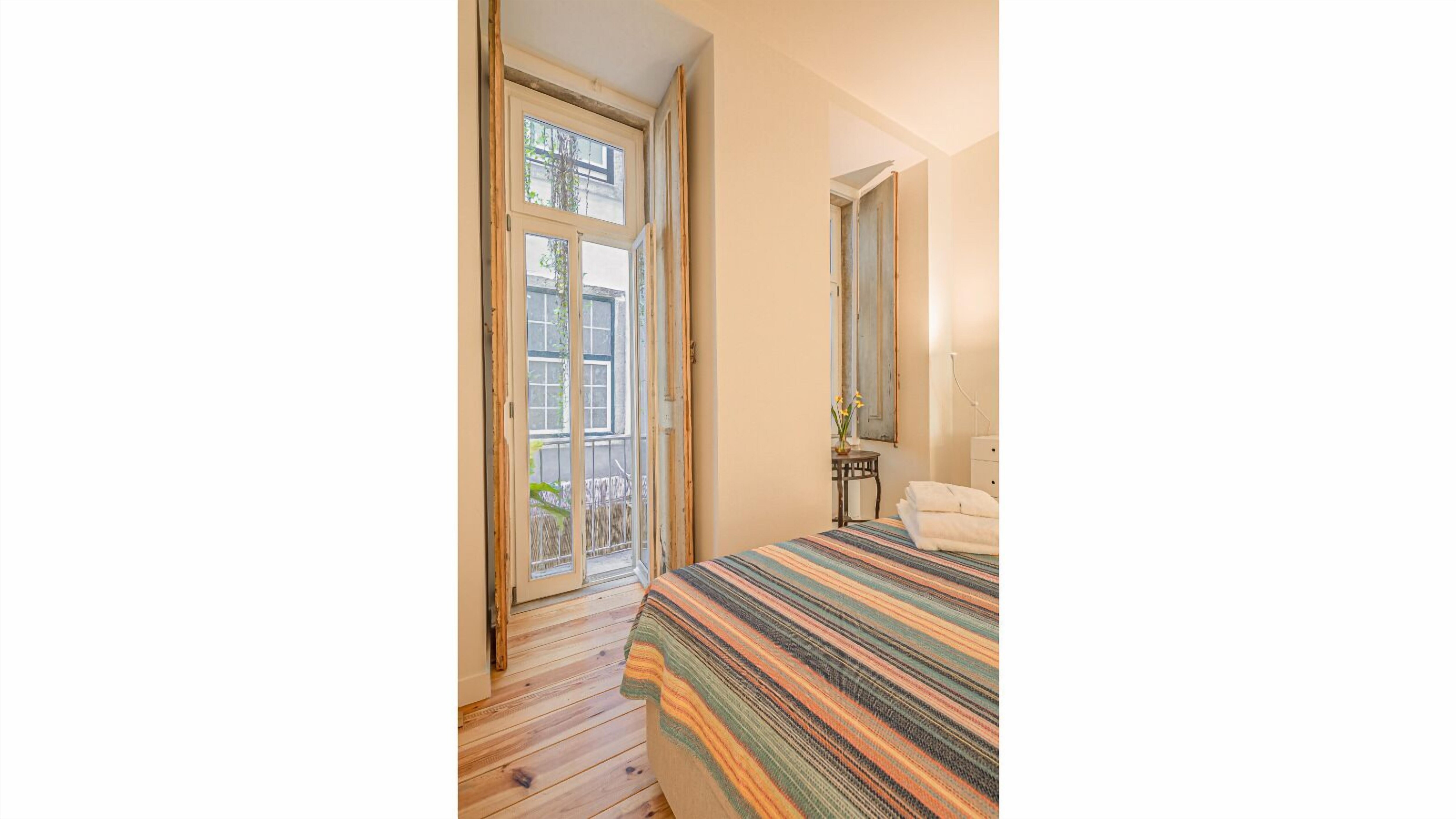 Una habitación con piso de madera, una cama con colcha a rayas, una mesita con un jarrón y un espejo en la pared.