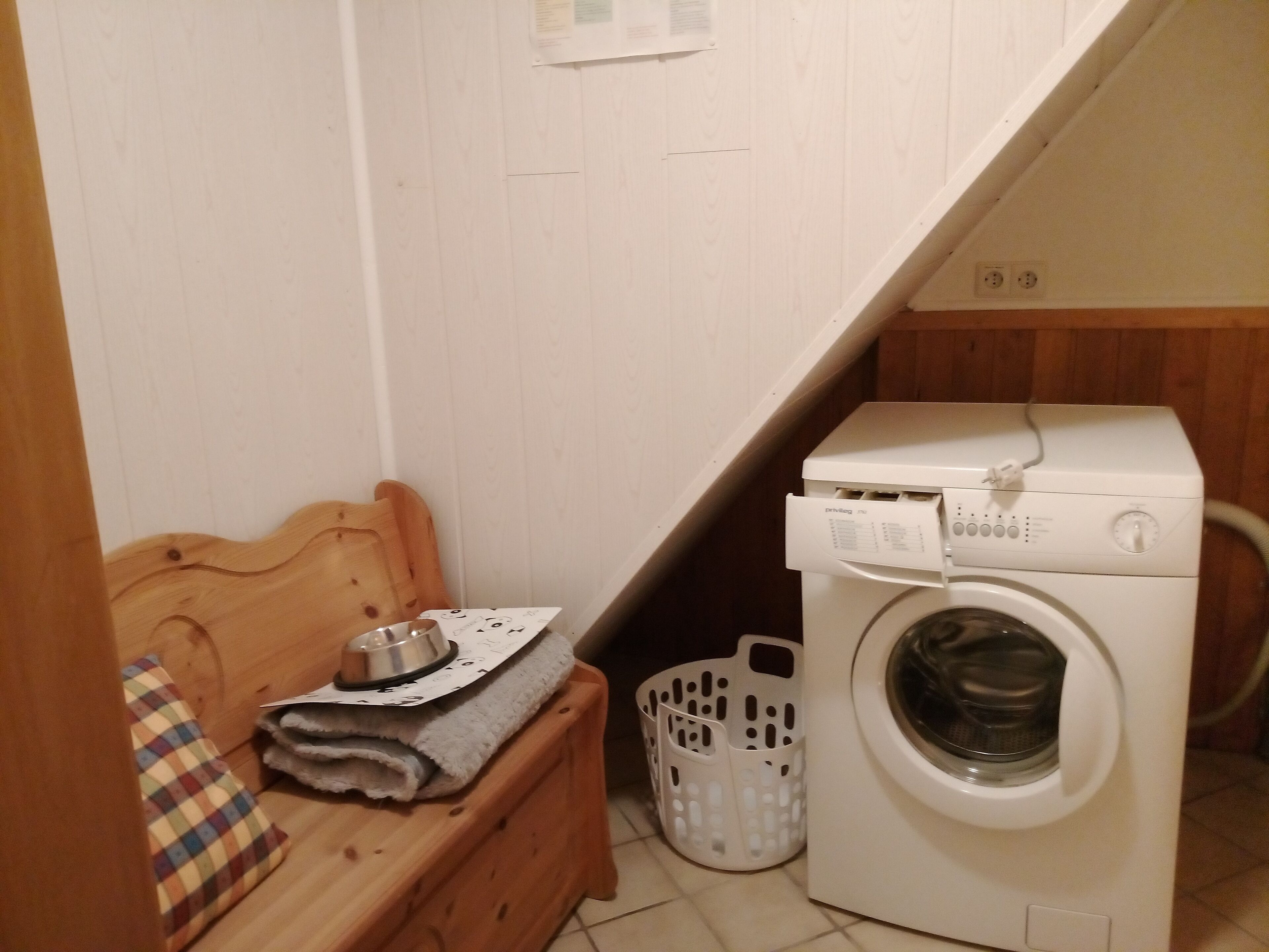 A laundry room with a washing machine, a wooden bench, a basket, and a towel rack.