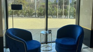 Una habitación moderna con dos sillones azules, una pequeña mesa redonda y ventanas amplias con vistas a edificios y árboles.