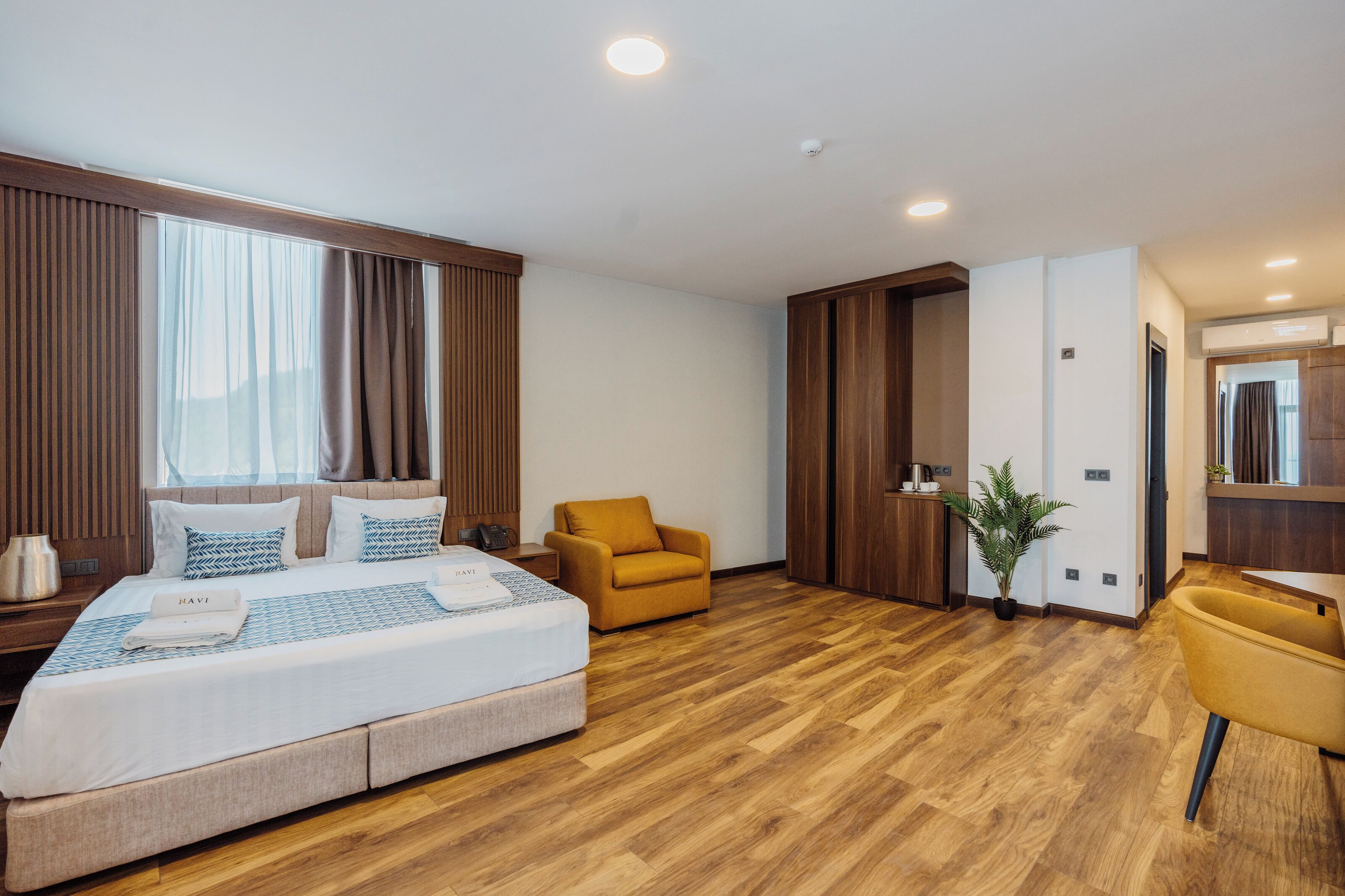 Una habitación de hotel moderna con una cama grande, un sillón amarillo, una mesita y una planta en maceta.