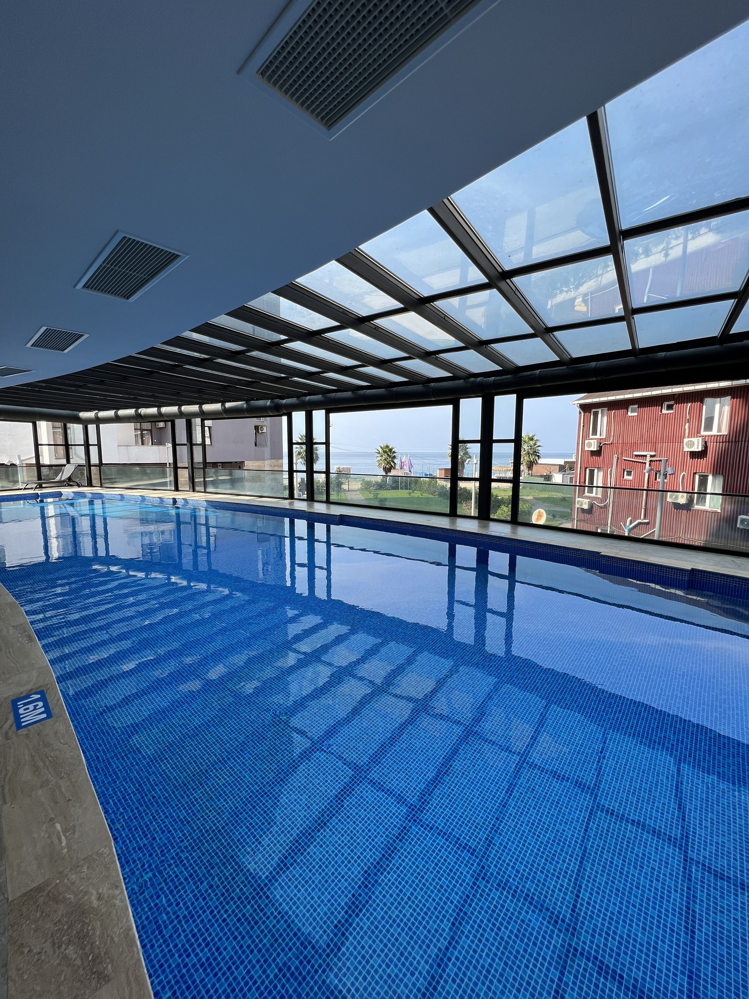 Une piscine intérieure à l’eau d’un bleu limpide, dotée de grandes fenêtres et offrant une vue sur un bâtiment rouge situé à l’extérieur.