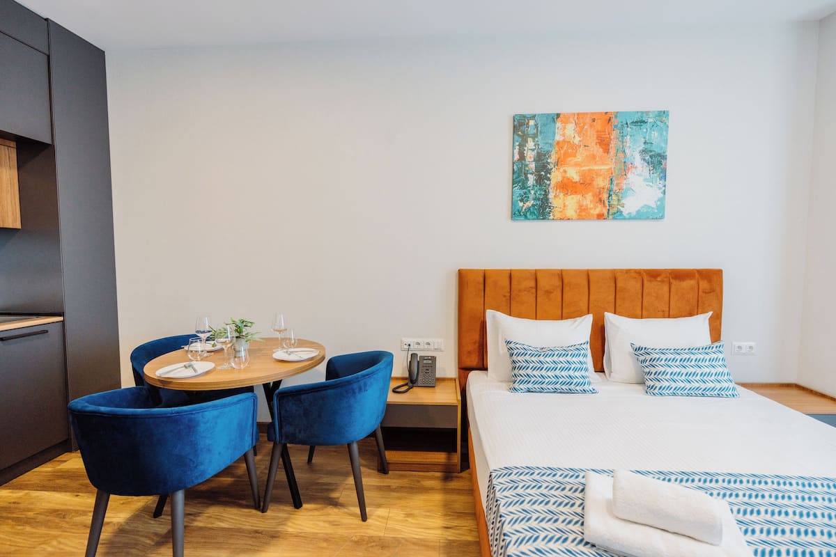 Quarto de hotel moderno com cabeceira de madeira, uma mesa de jantar redonda com cadeiras azuis e uma cama com roupa de cama estampada.