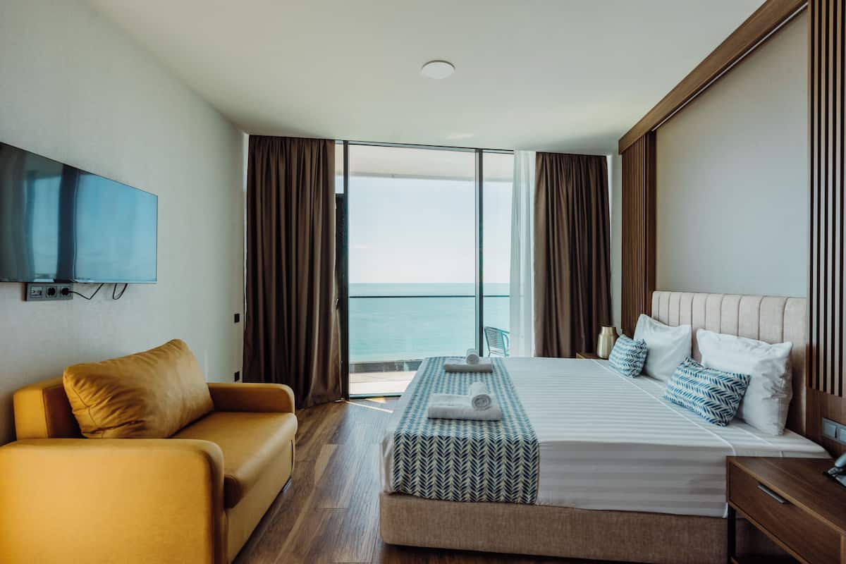 Quarto de hotel com uma cama grande, um sofá, uma televisão na parede e vista para o oceano pela porta de correr de vidro.