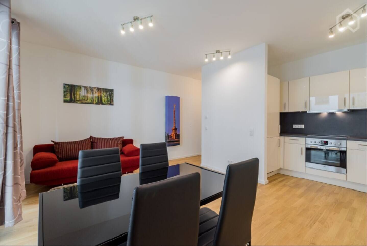 Una sala moderna con un sofá rojo, una mesa de vidrio y sillas negras. Cuenta con una zona de cocina con armarios blancos y una estufa.