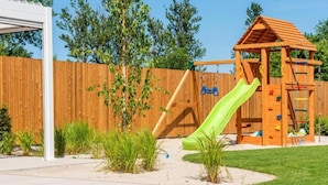 A wooden play structure with a slide and climbing area, surrounded by a wooden fence and greenery. - Comfortable, convenient holiday apartment (Grzybowo)