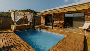 Une piscine avec une terrasse en bois, un espace salon couvert et un bâtiment en briques doté de grandes fenêtres.
