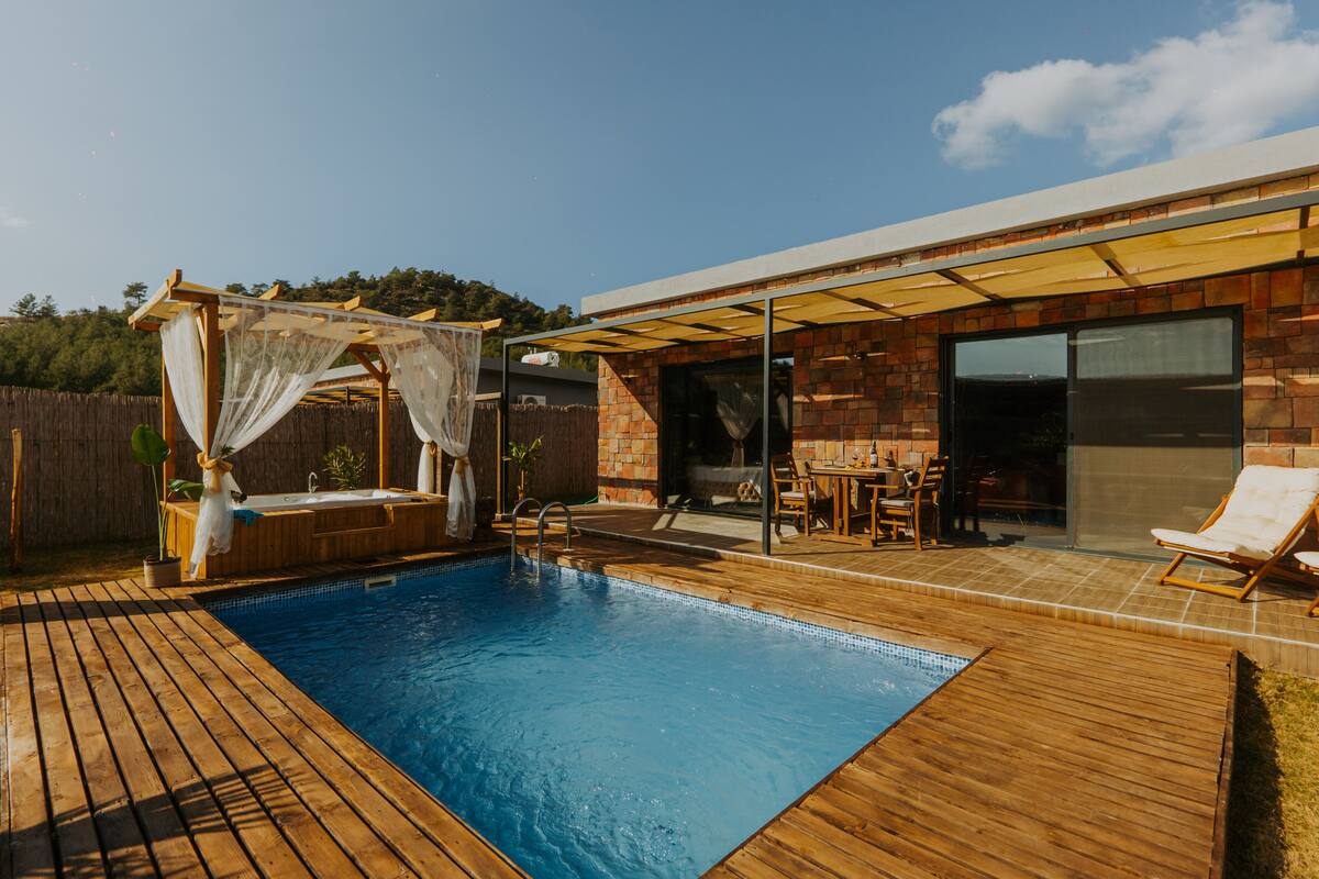 A swimming pool with a wooden deck, a covered seating area, and a brick building with large windows.