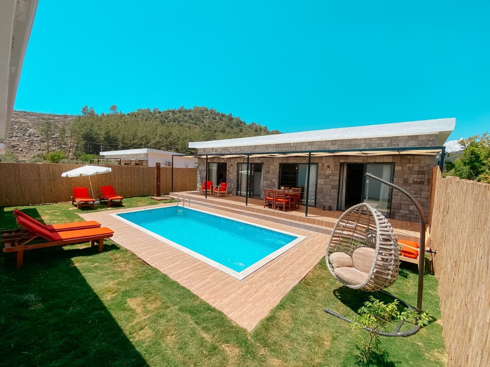 A modern outdoor pool area with a wooden deck, red lounge chairs, and a hanging chair.