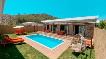 A modern outdoor pool area with a wooden deck, red lounge chairs, and a hanging chair.