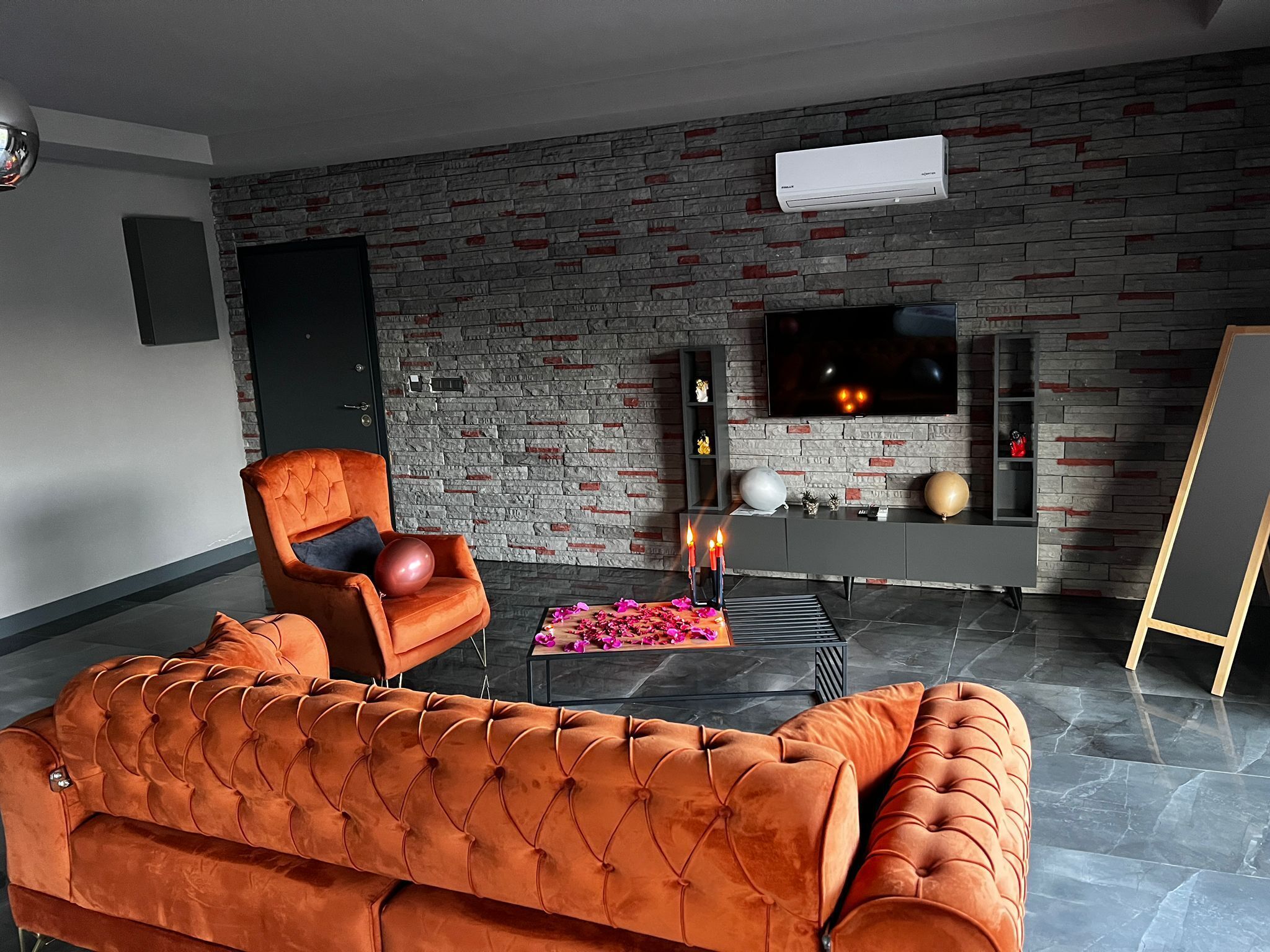 A modern living room with a brick wall, a sofa, a chair, a coffee table with flowers, a TV, and a fireplace.