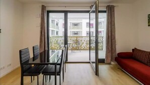 Un salon comprenant une table à manger et des chaises, un canapé rouge et une porte-fenêtre coulissante donnant sur un balcon avec vue sur d’autres immeubles.