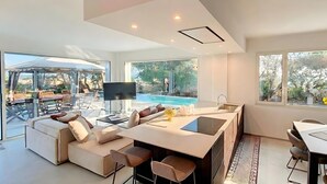 A modern living room with a large sofa, a dining table, and a kitchen island. There is a pool visible through the large windows. - 5-star dream villa, beach access, pool, jacuzzi (Marina di Felloniche)