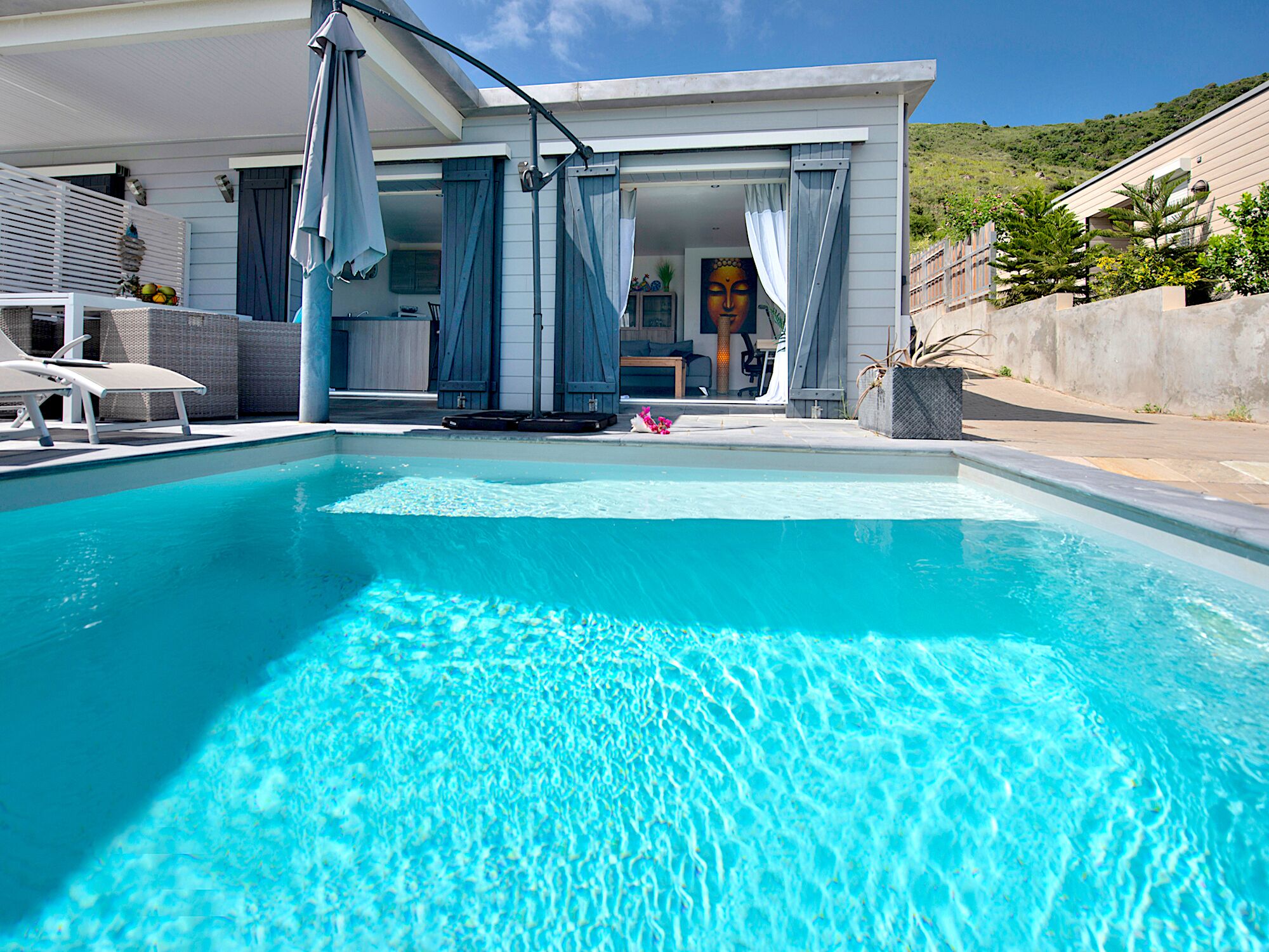 A modern pool area with clear blue water, a covered seating area with umbrellas, and a building with large windows and curtains.