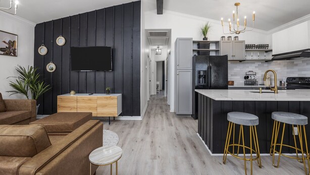 Una cocina moderna con una isla central, taburetes de bar, un televisor de pantalla plana y una zona de comedor con un sofá marrón y una pequeña mesa redonda.