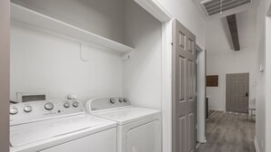 Un lavadero con dos lavadoras y secadoras de carga frontal, un respiradero en el techo blanco y un pasillo que conduce a otra habitación.