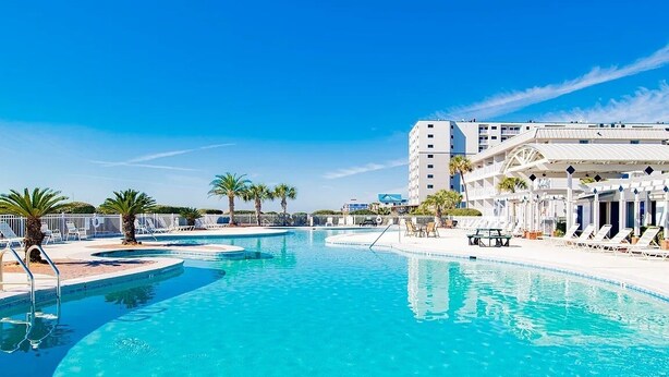 Una amplia piscina con tumbonas y sombrillas, rodeada de palmeras y un edificio de hotel.
